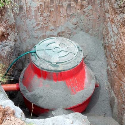 Red septic tank in a dirt trench, covered with a green lid, surrounded by gray sand, and a pipe.