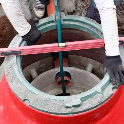 Workers installing sewer manhole, using a level. Concrete and red casing visible.