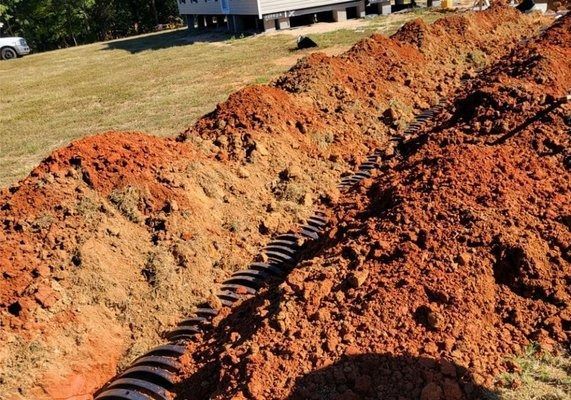 Trench dug in red soil, holding a black corrugated pipe. Pile of dirt beside the trench, green grass in the background.