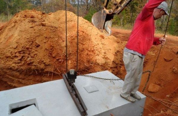 Man using ropes to guide concrete septic tank installation near a dirt pile and excavator.
