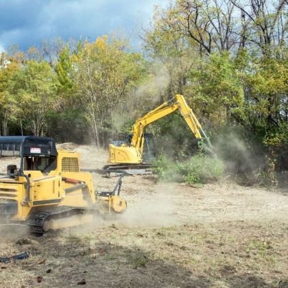 Yellow construction vehicles clearing brush and trees in a field; trees in background.