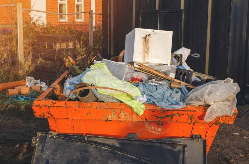 Orange dumpster overflowing with trash at a construction site; brick building in the background.