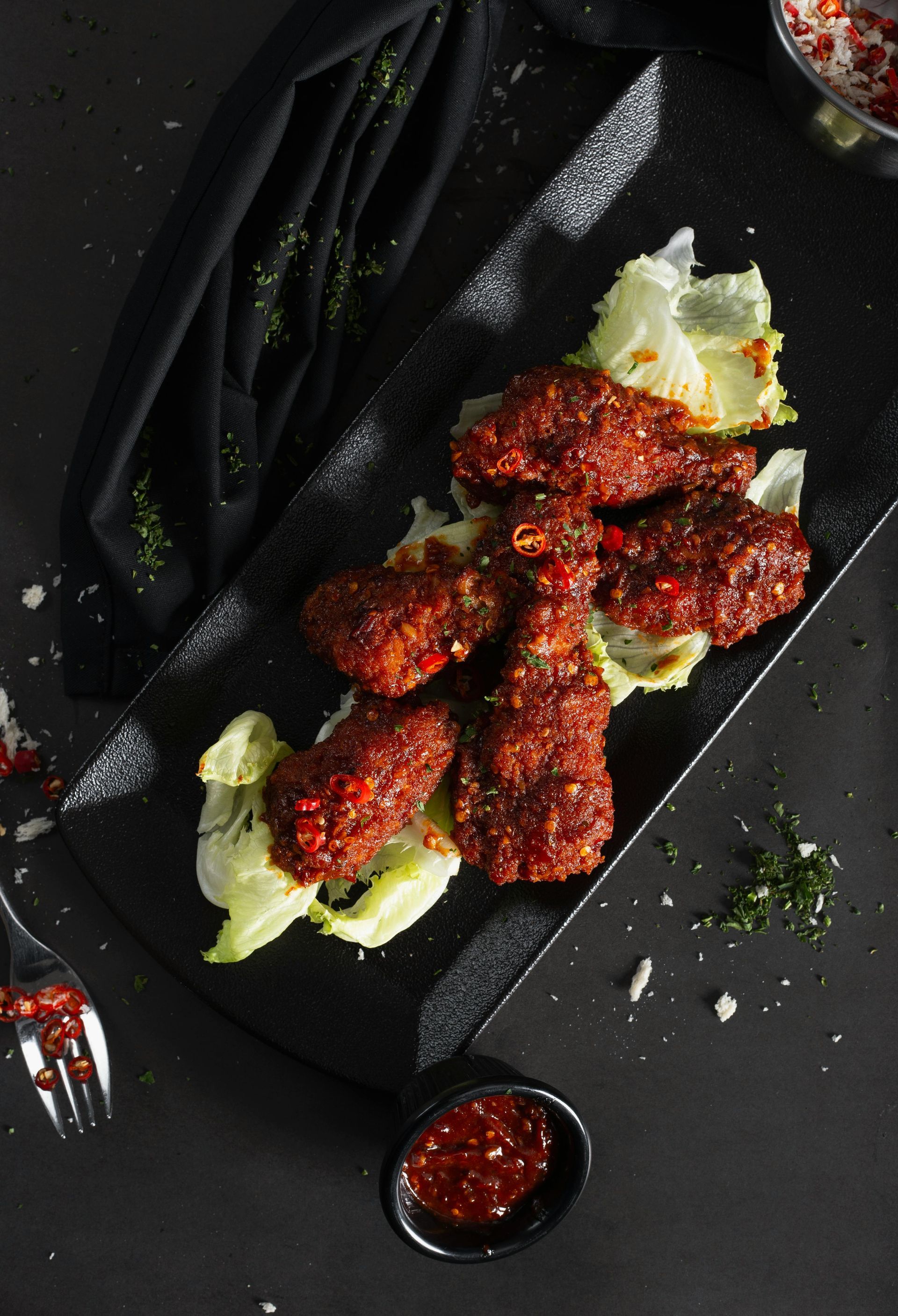 Spicy fried chicken wings on a black plate with lettuce and a small bowl of sauce.