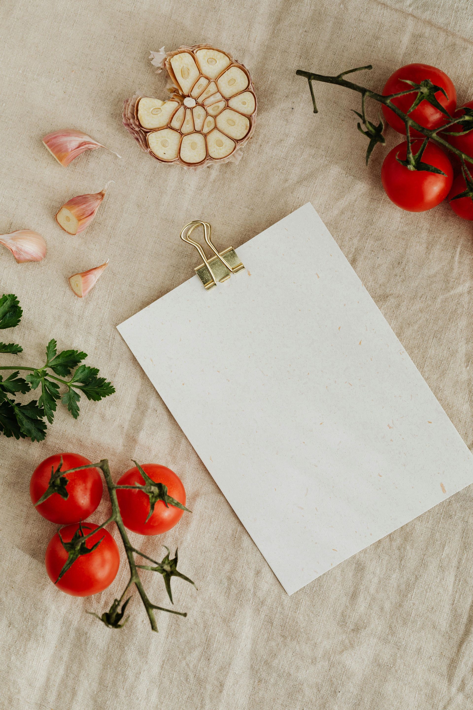 Tomatoes, garlic, parsley, and blank paper clipped together on a textured beige surface.