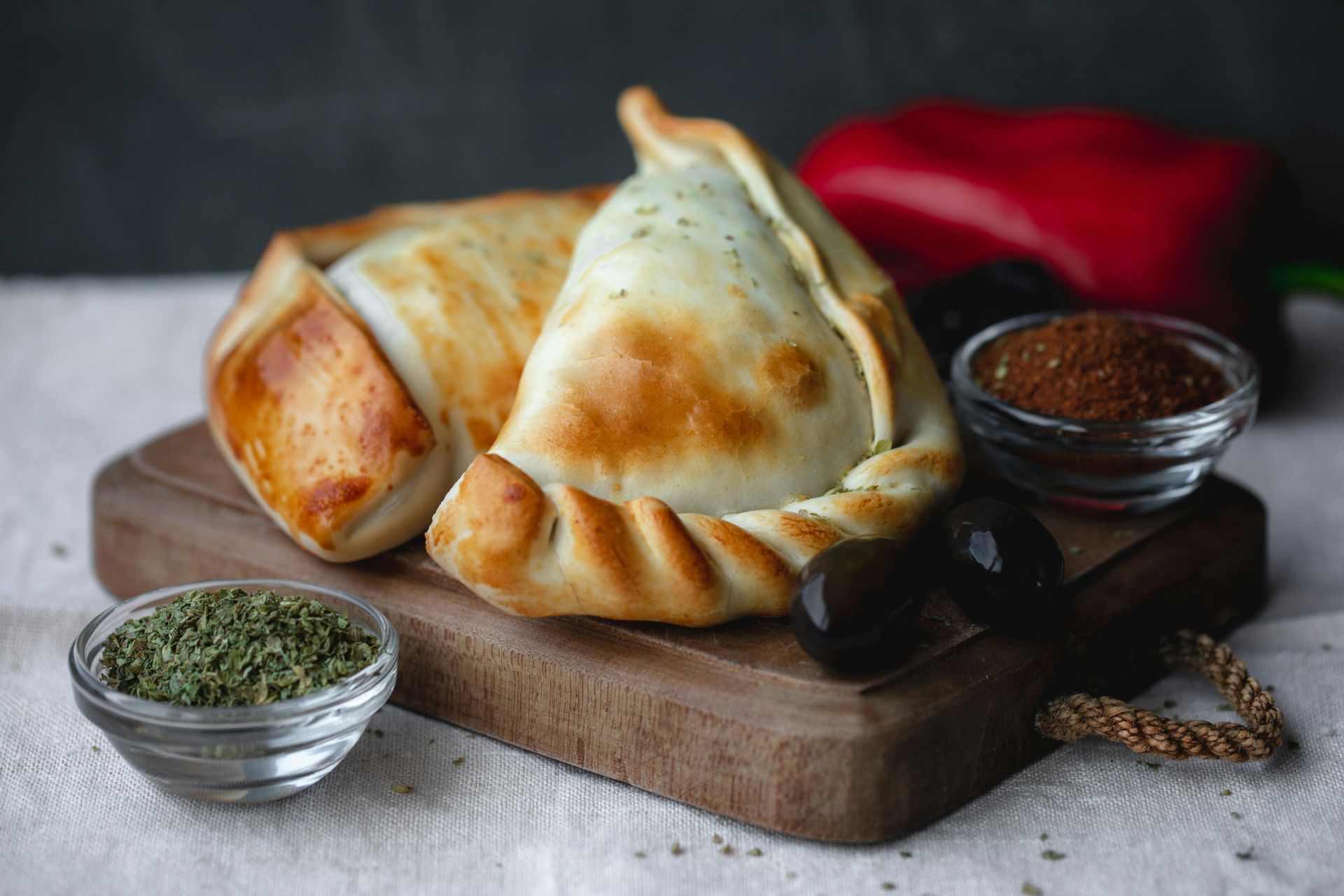 Two baked empanadas on a wooden board, with herbs, olives, and spices.