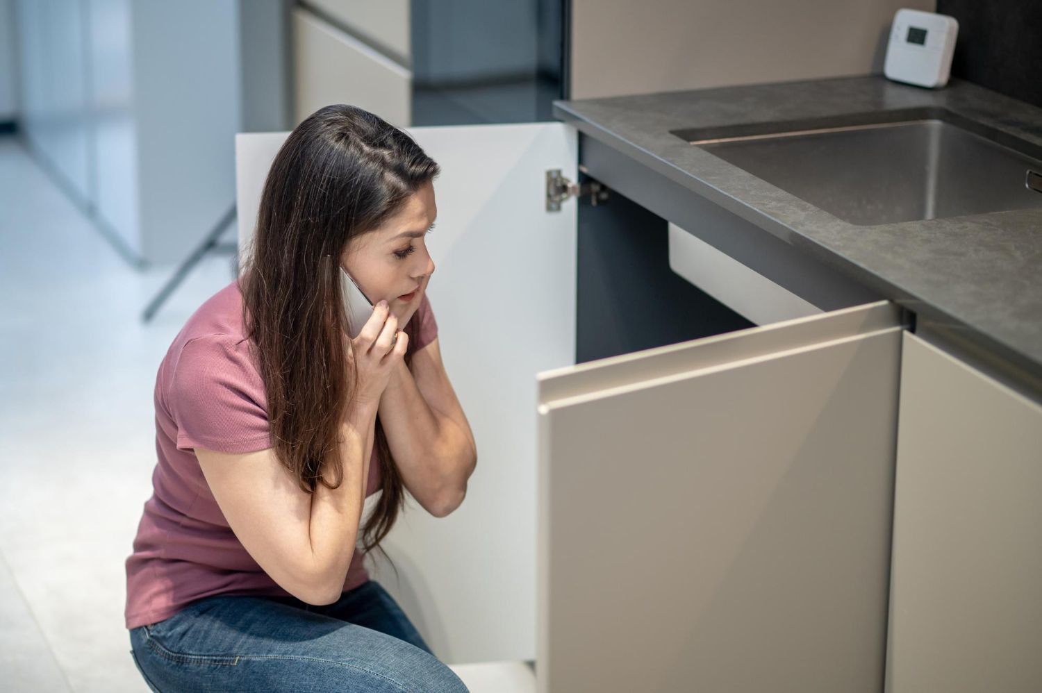 A woman is sitting on the floor in a kitchen talking on a cell phone.