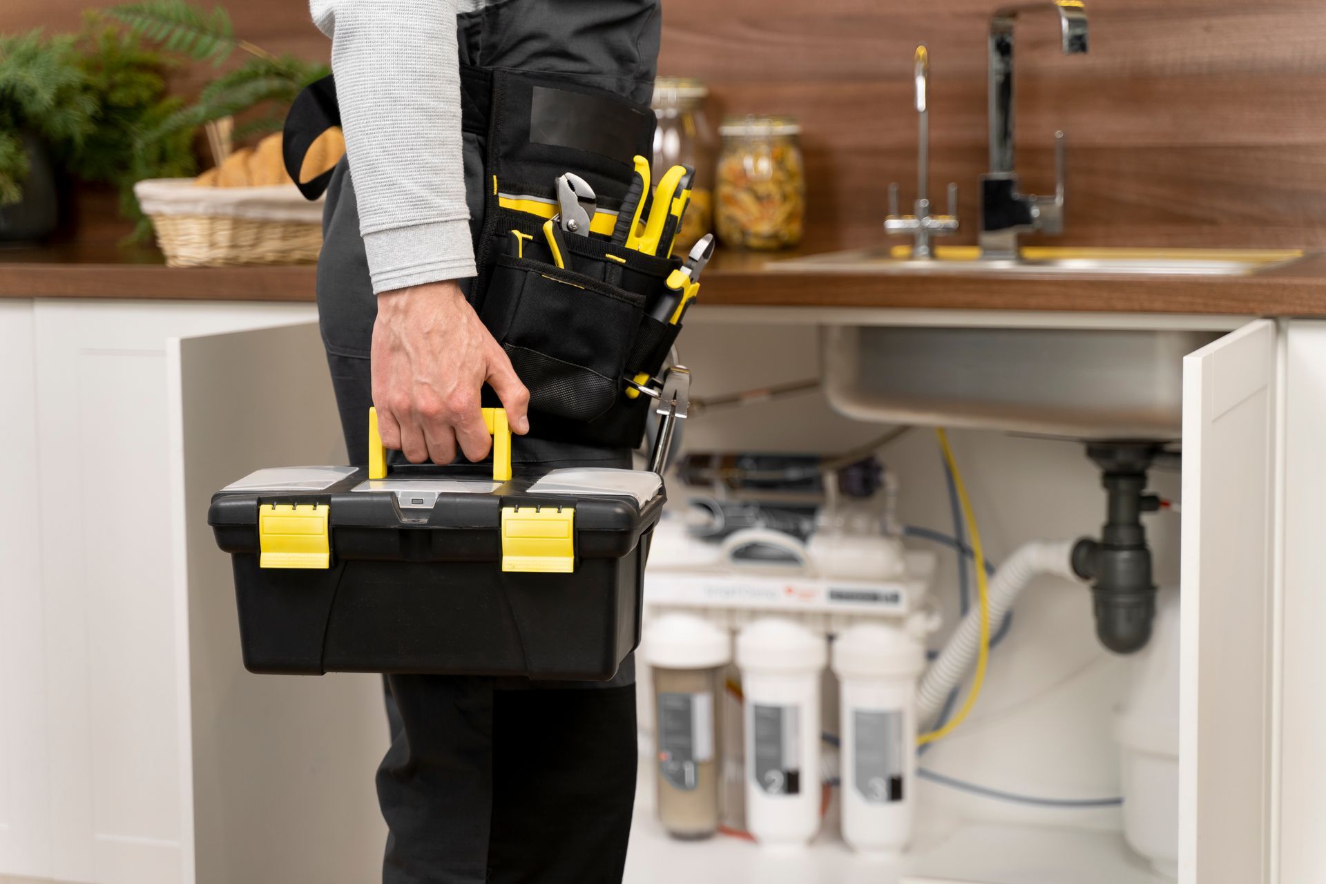 A plumber is holding a toolbox in front of a sink in a kitchen.