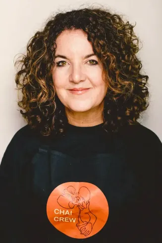 A woman with curly hair is wearing a black shirt with a sticker on it that says chai crew.