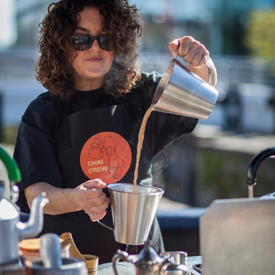 A woman wearing a shirt that says chai crew is pouring coffee into a cup