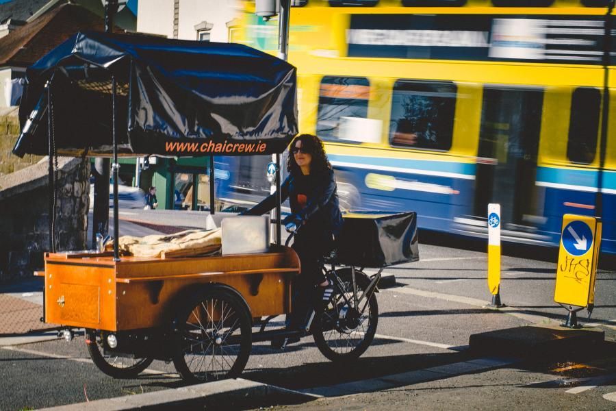 A woman is riding a bike next to a cart that says ' www.chatbrew.ie ' on it