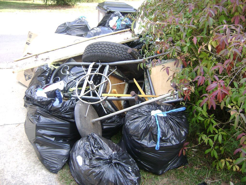 Pile of black trash bags, old tires, and household junk on a driveway.