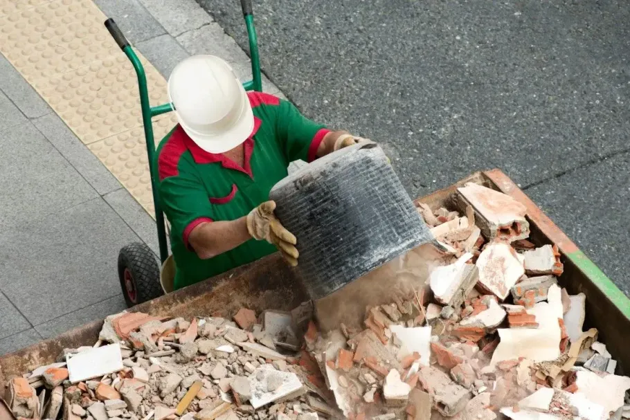 Worker in a hard hat dumping renovation debris into a large disposal bin.