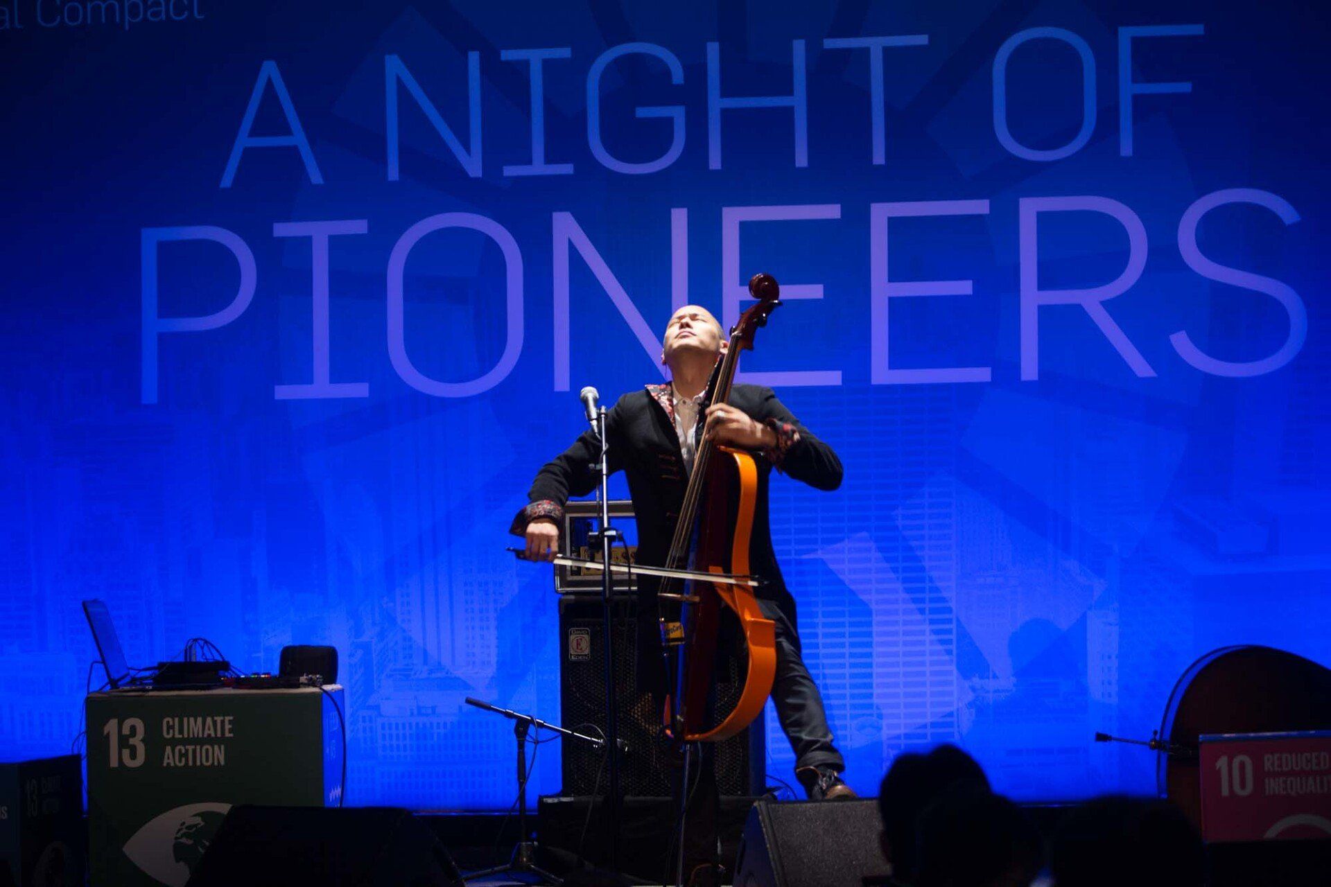 A man playing a cello in front of a sign that says a night of pioneers