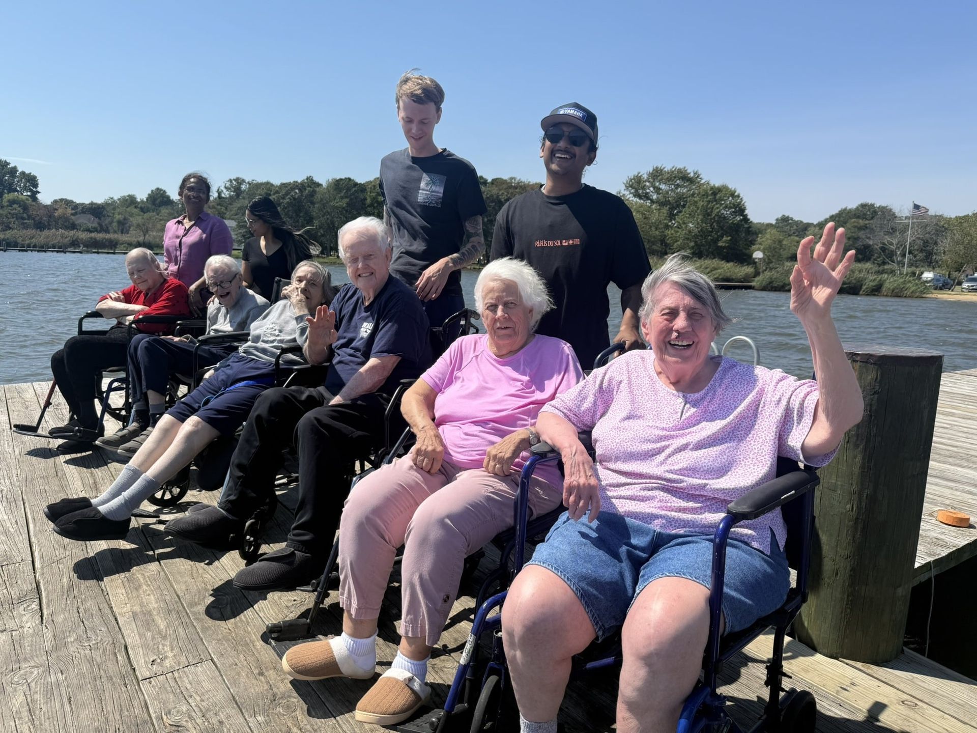 A group of people in wheelchairs and standing individuals on a wooden dock outdoors near a body of water.