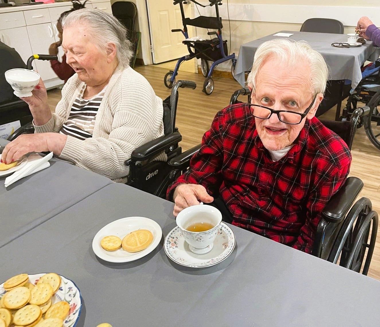 Two people in wheelchairs sit at a table with tea and cookies, one smiling at the camera and the other drinking from a cup.
