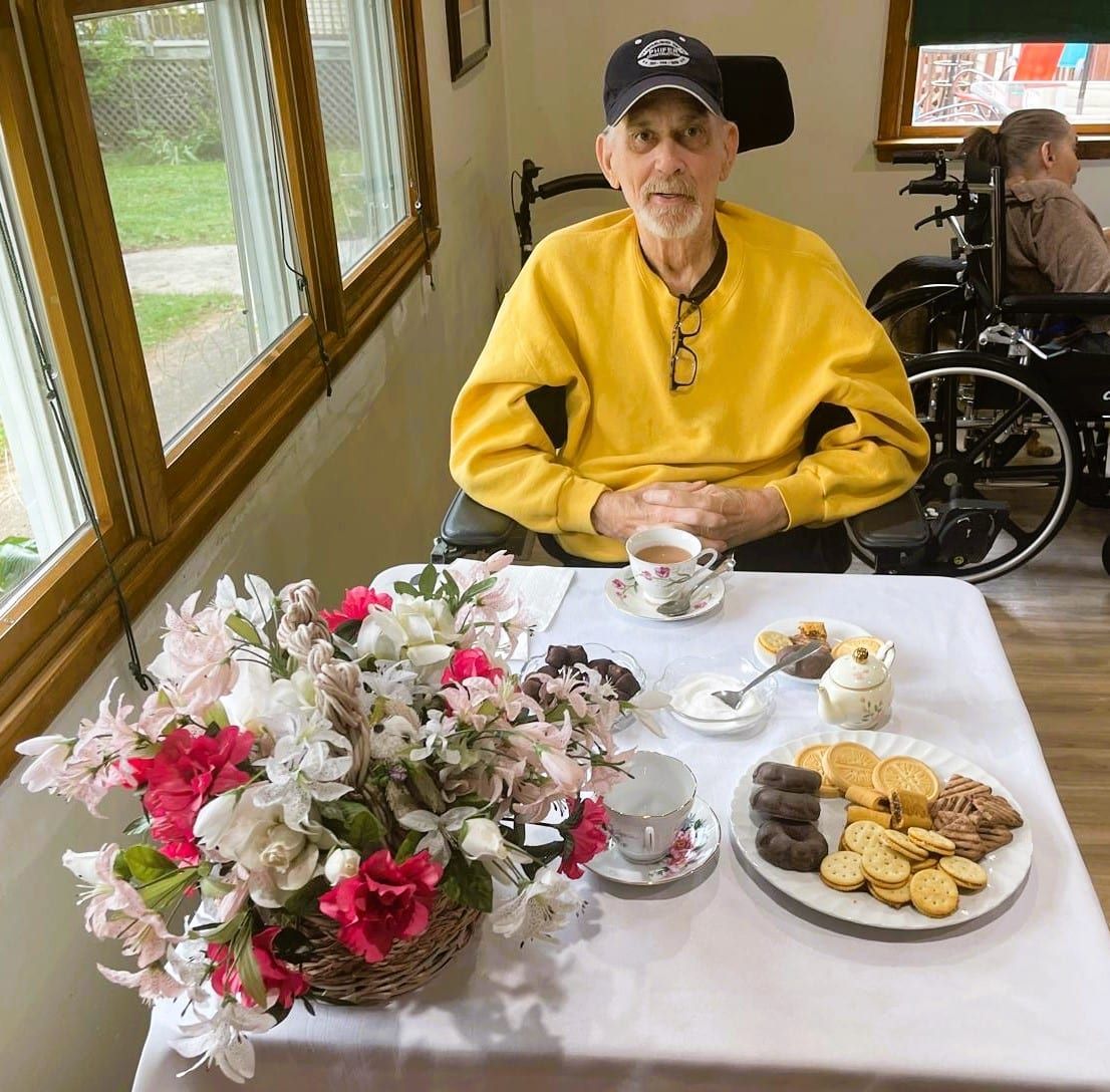 A person in a yellow sweater sits at a table with a flower arrangement, tea cups, and a plate of snacks by a window.