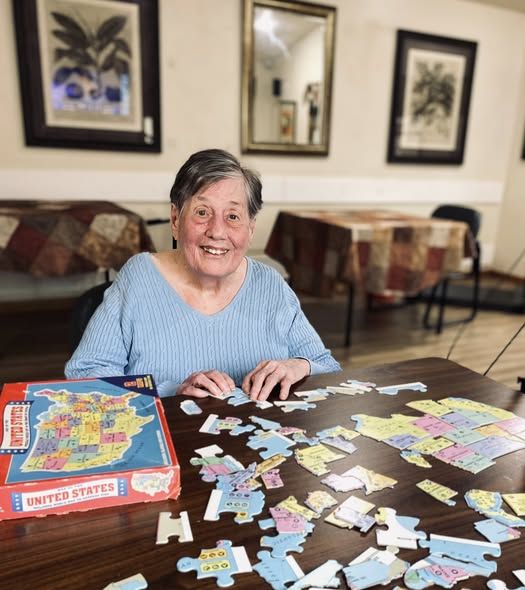 A smiling person assembling a United States map puzzle at a wooden table in a room with framed art and covered tables.