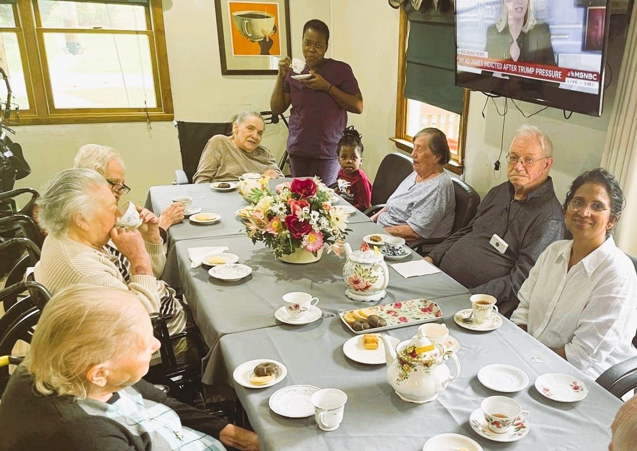 A group of people sit around a long table with tea and refreshments, with a staff member serving and a television visible.