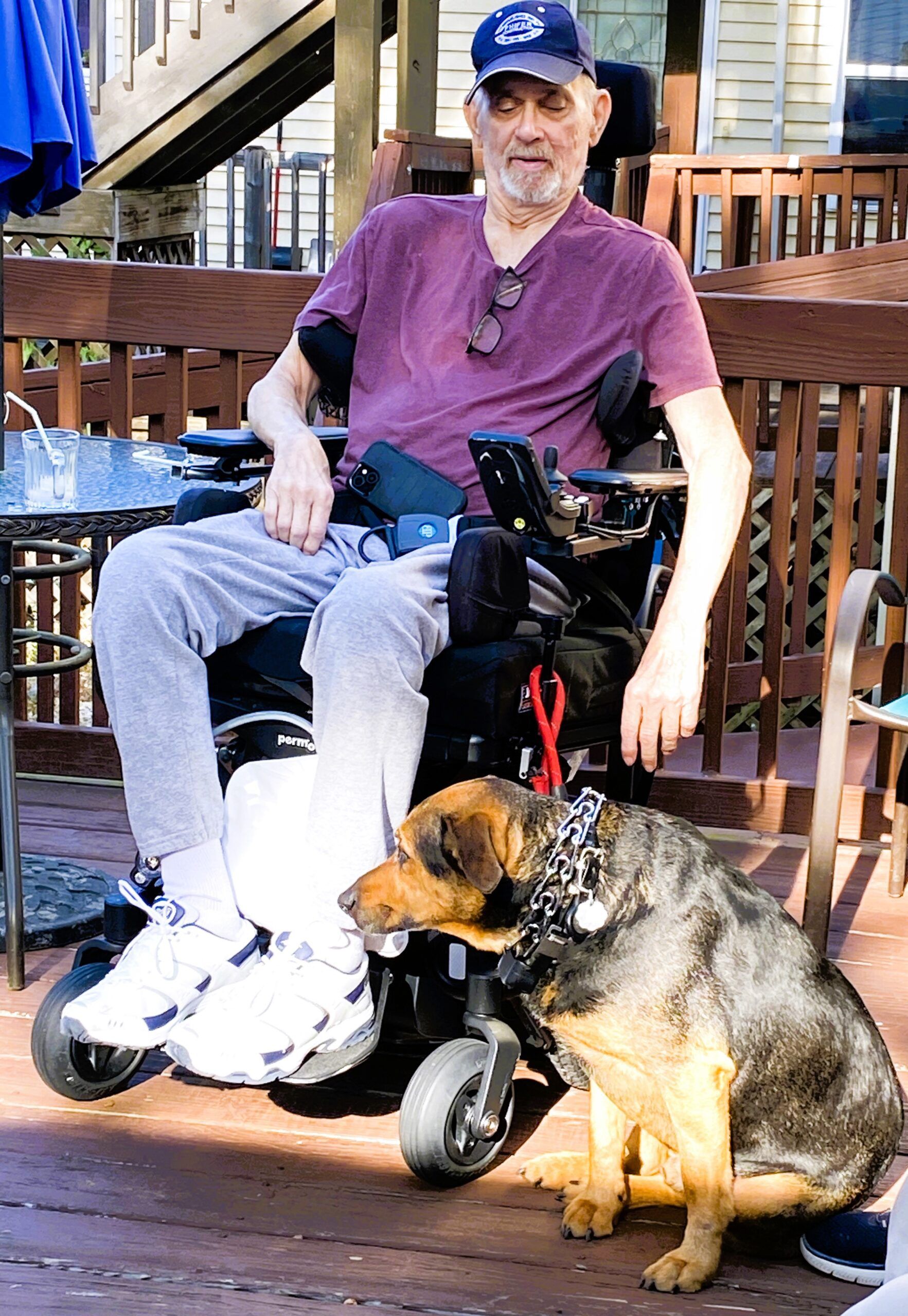 A person in a motorized wheelchair sits on a wooden deck next to a medium-sized dog with a patterned collar.
