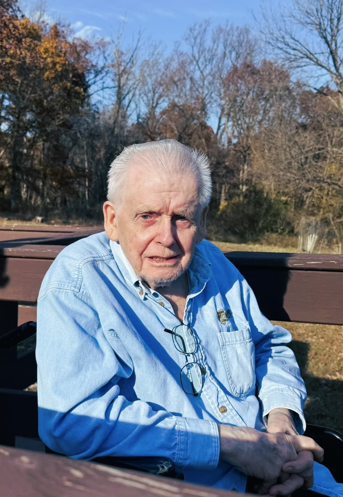 A person in a blue button-down shirt with glasses hanging from their collar, sitting outdoors during a sunny day.