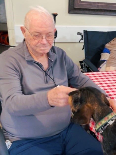 An elderly man in a gray sweater gently pets a small, dark dog at a table with a red-and-white checkered tablecloth.
