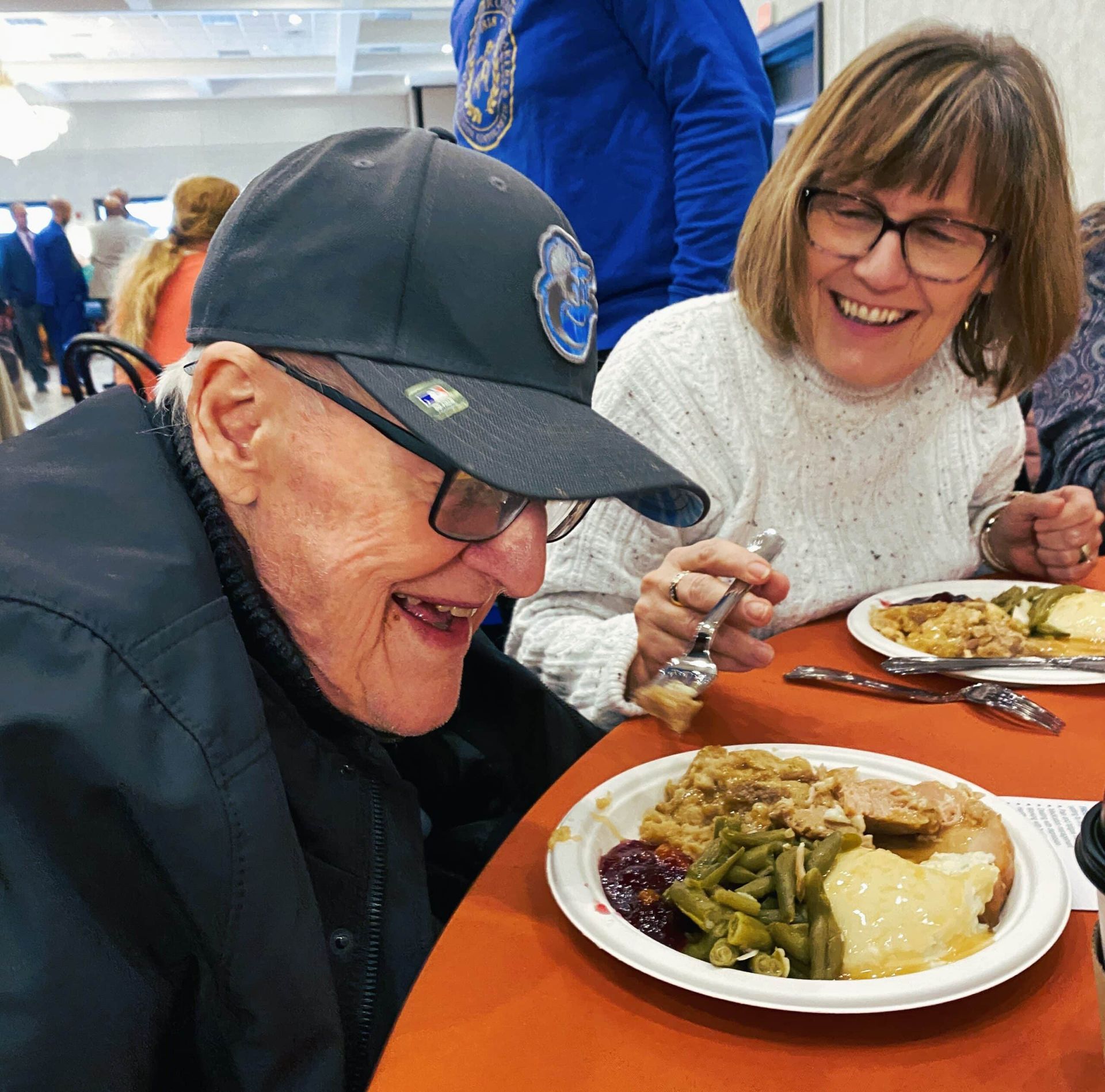 Two people smiling while seated at a table with plates of food during a gathering.