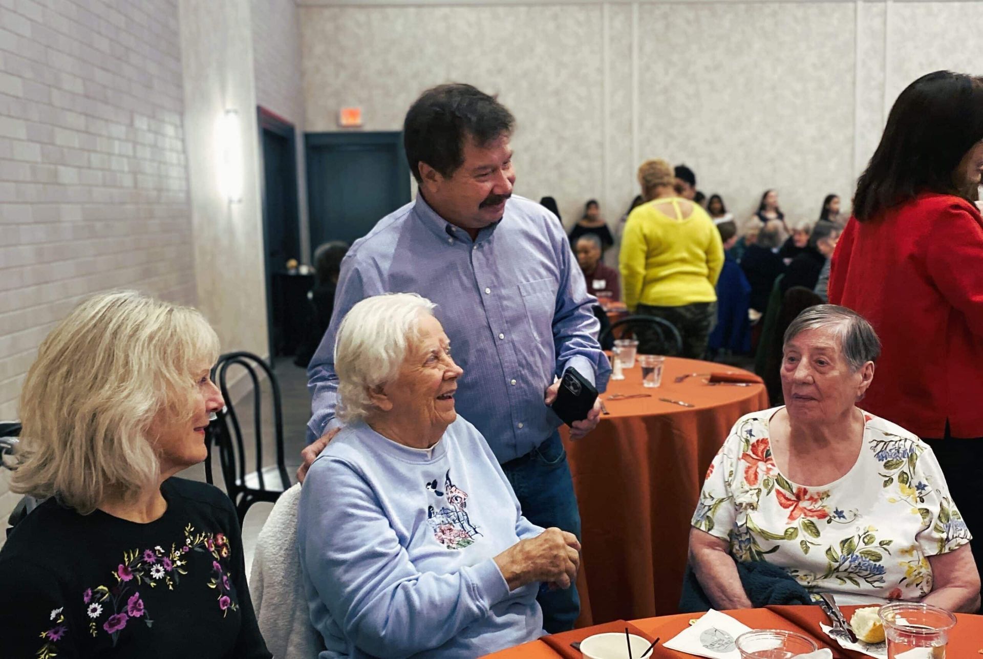 A person in a light blue shirt stands at a table, talking with two seated people in a room with orange tablecloths.