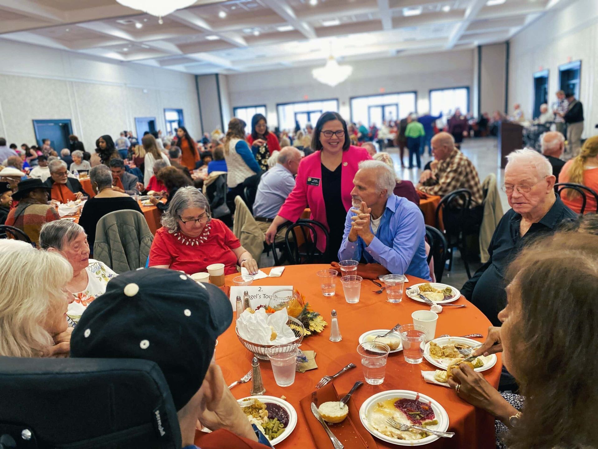 People sit at round tables with orange tablecloths in a large hall during an event, some talking and others eating.