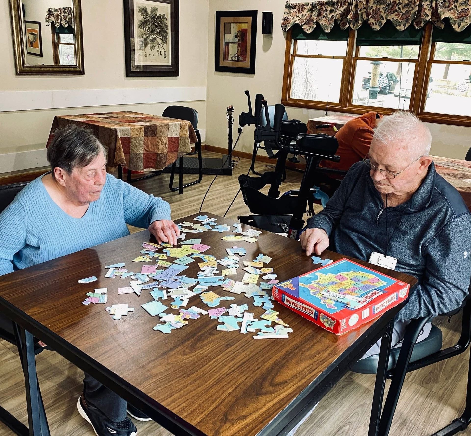 Two people sit at a wooden table in a room, working together on a jigsaw puzzle with a colorful box nearby.