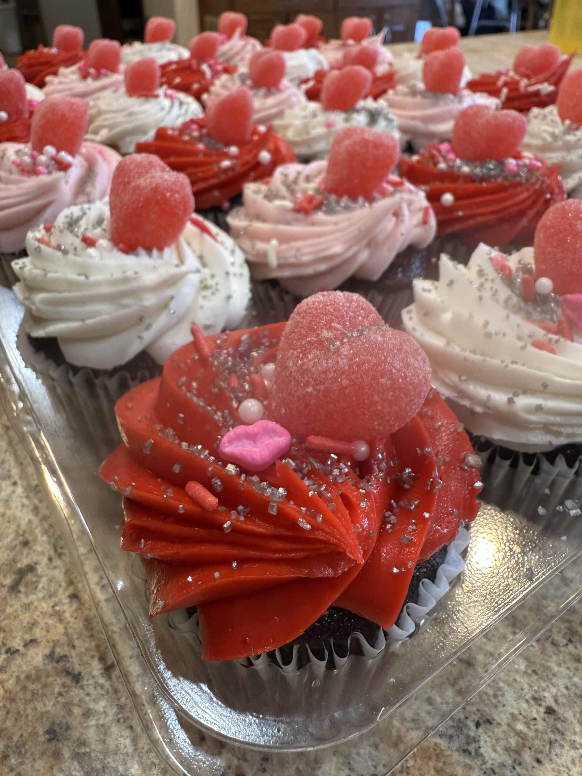 A close-up of Valentine's Day cupcakes with red and pink frosting, decorated with gummy hearts and silver sprinkles.