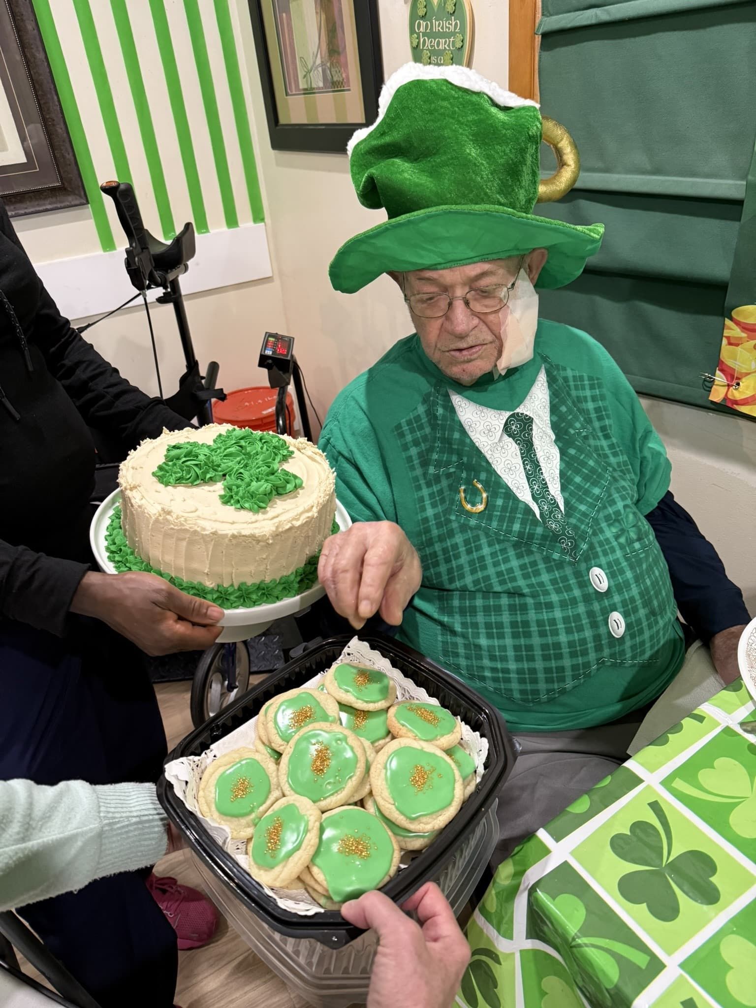 A person in a green leprechaun hat and outfit reaches for a platter of green frosted cookies near a St. Patrick's cake.