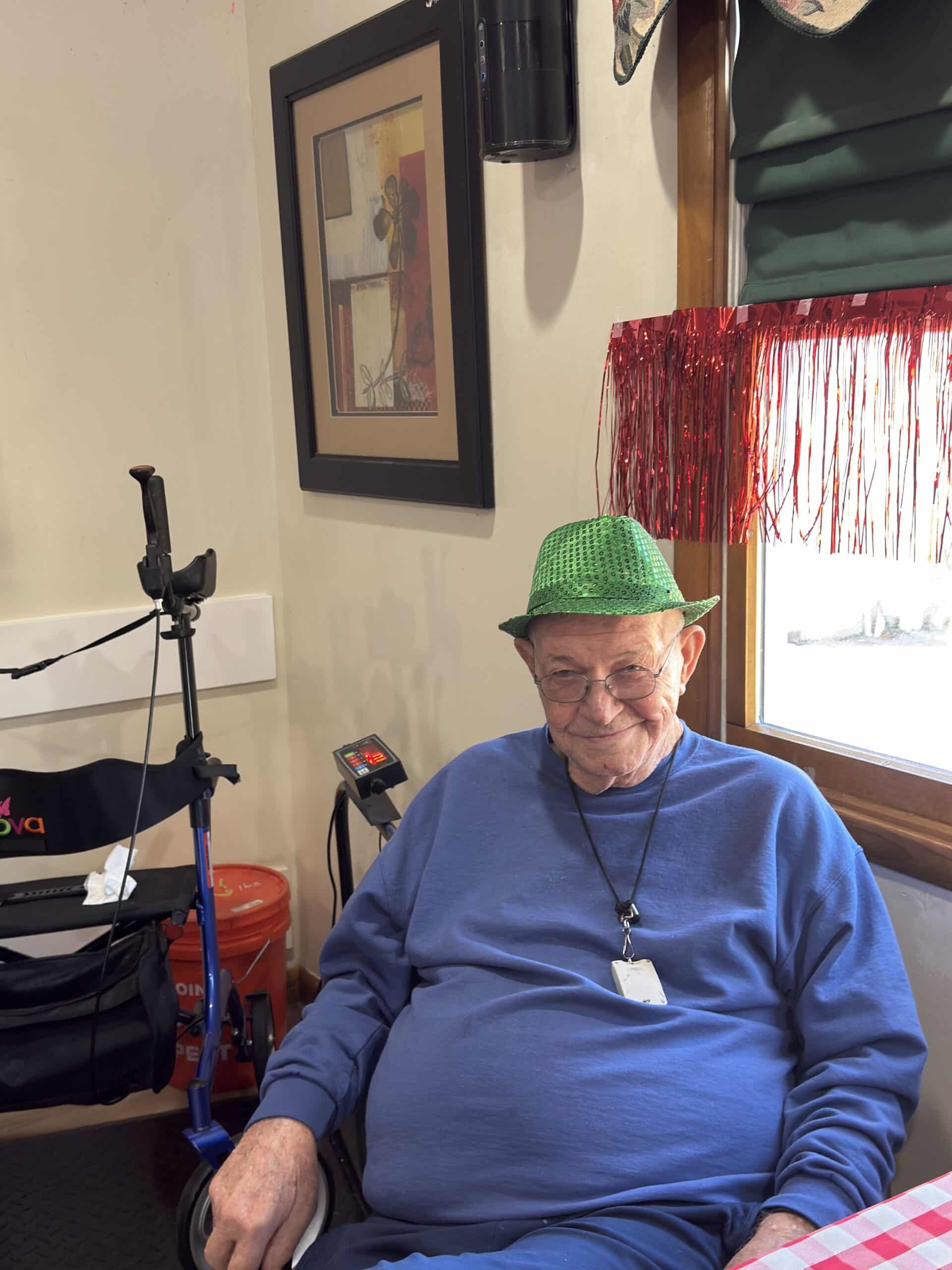 A smiling person wears a green patterned hat and a blue long-sleeved shirt, seated near a walker and red fringe curtains.