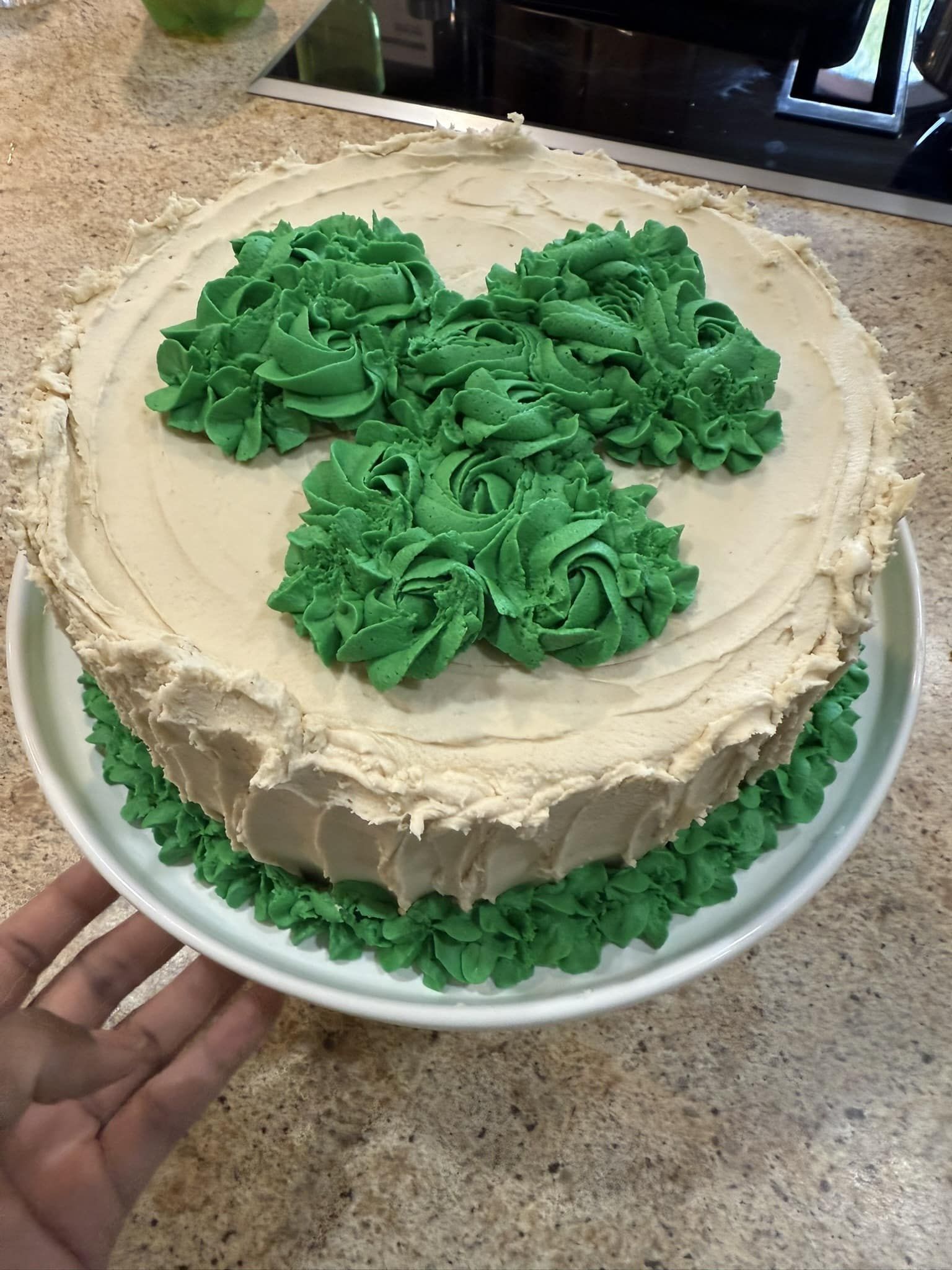 A round cake with beige frosting, decorated with three green icing rosettes on top and a green piped border at the base.