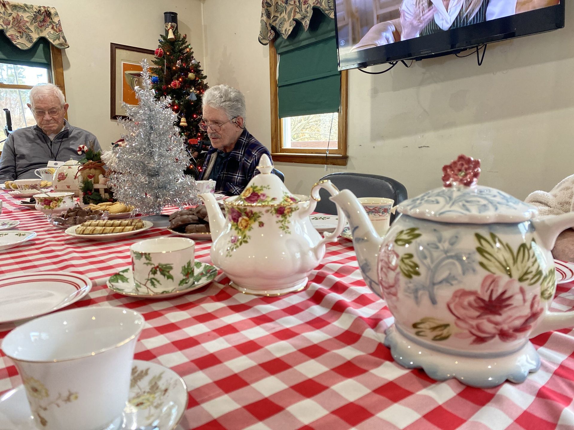 A table set for tea with patterned porcelain teapots and cups on a red gingham tablecloth, with two people in the back.