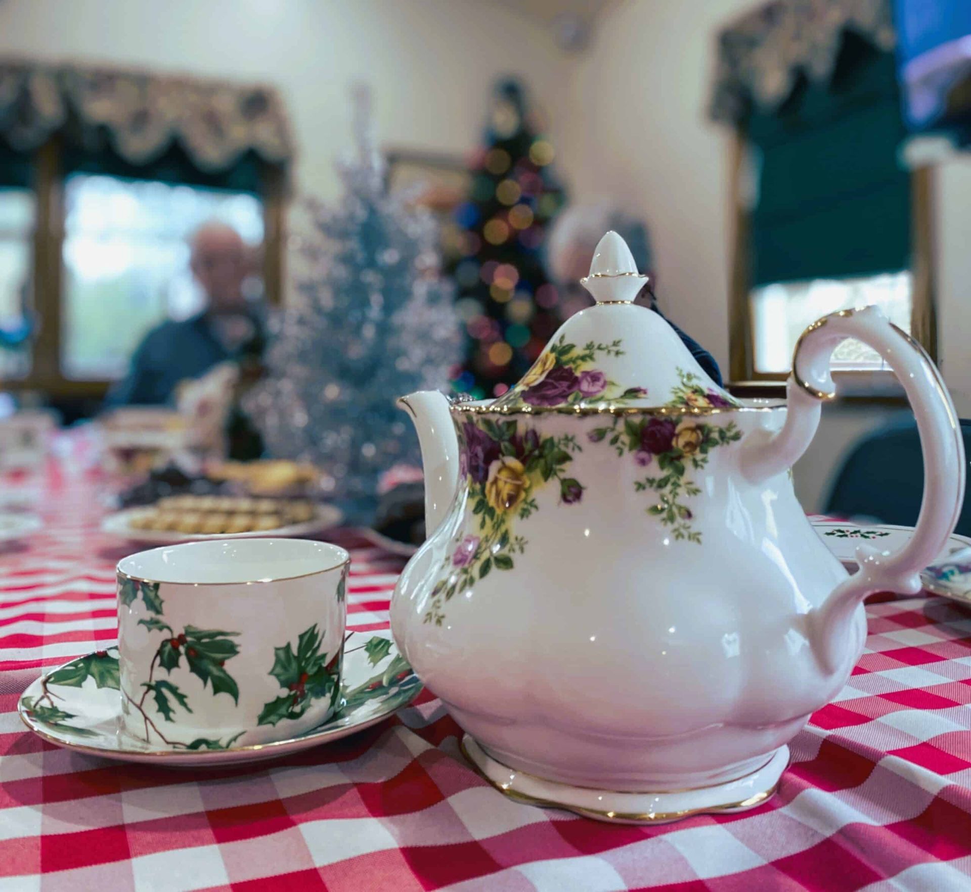 A floral teapot and matching teacup on a red-and-white checkered tablecloth, with a blurred holiday scene in the back.