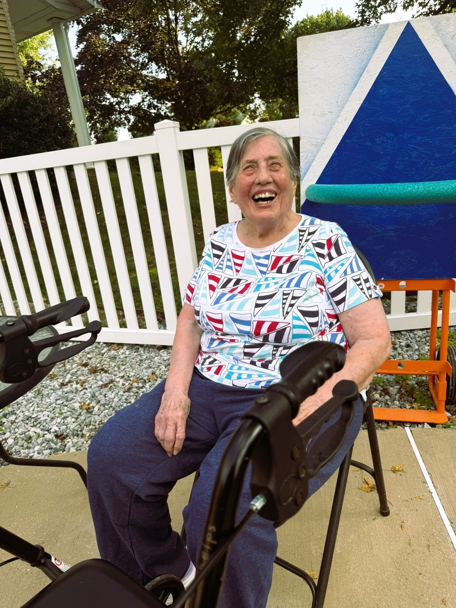 A smiling individual sits on a walker on a patio with a white fence and a blue triangular sign in the background.