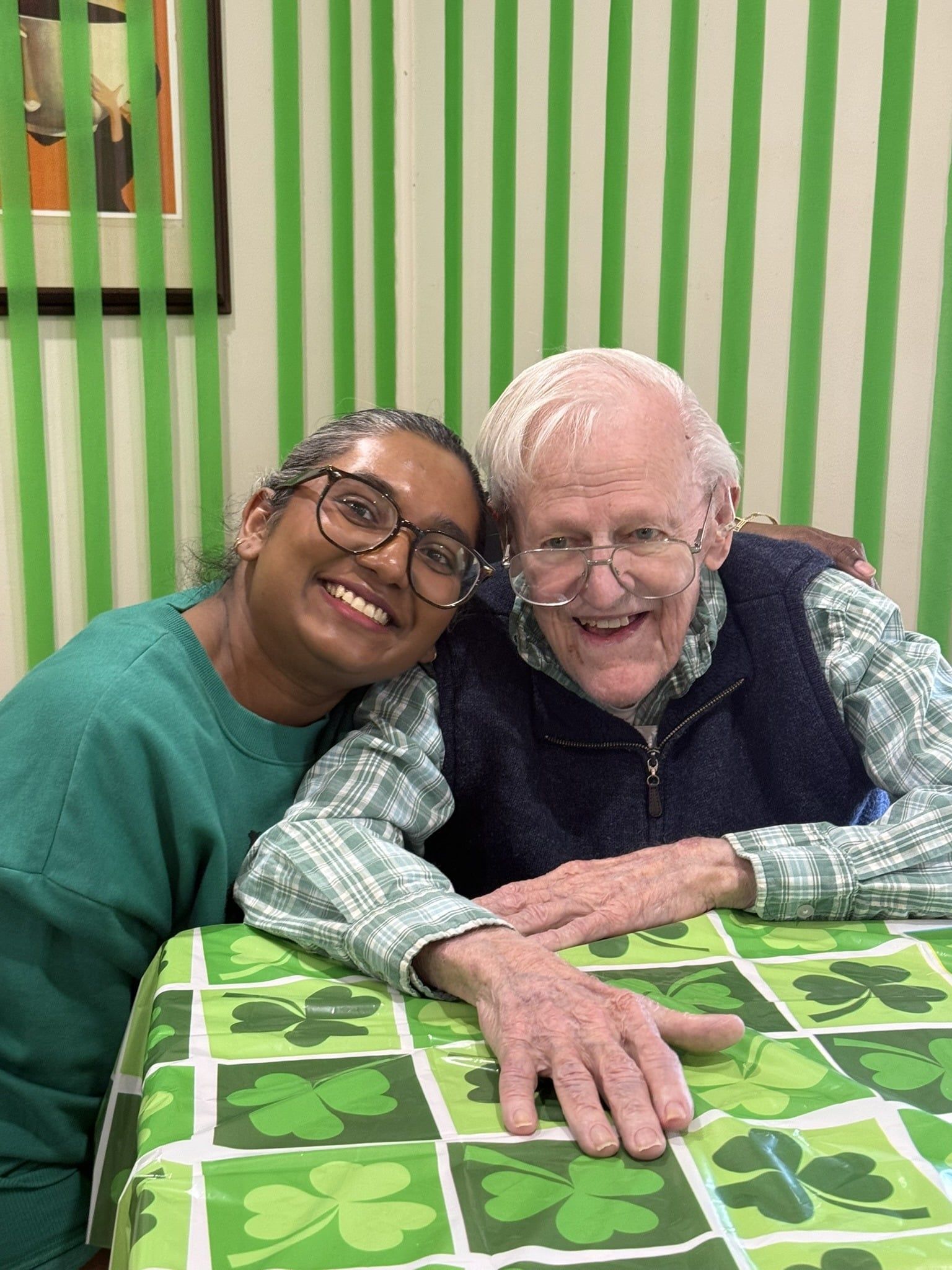 Two people sit together at a table with a green shamrock cloth, smiling for a photo against a green-striped backdrop.