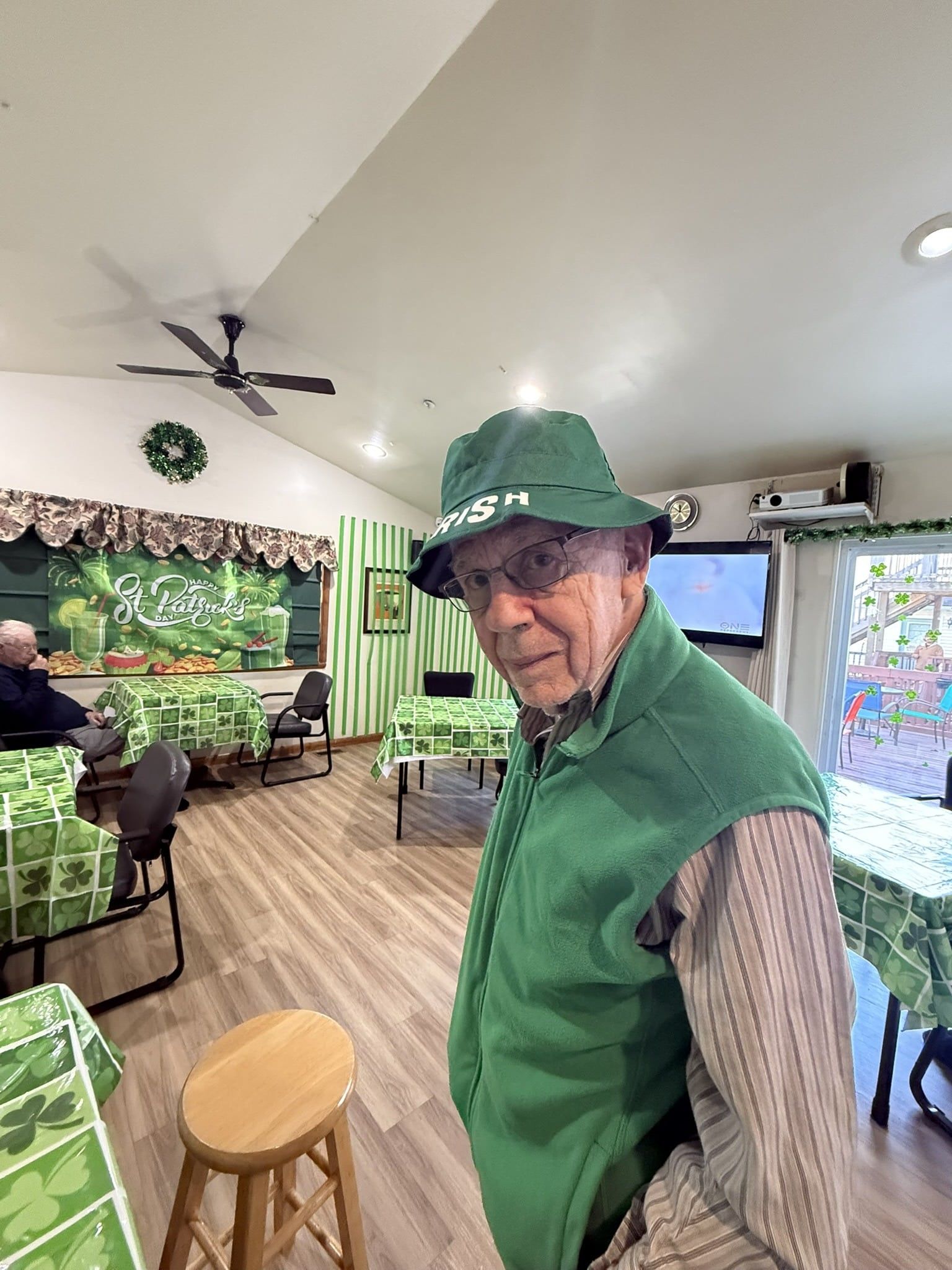 A person in a green hat and vest stands in a room decorated with shamrock-patterned tablecloths and green curtains.