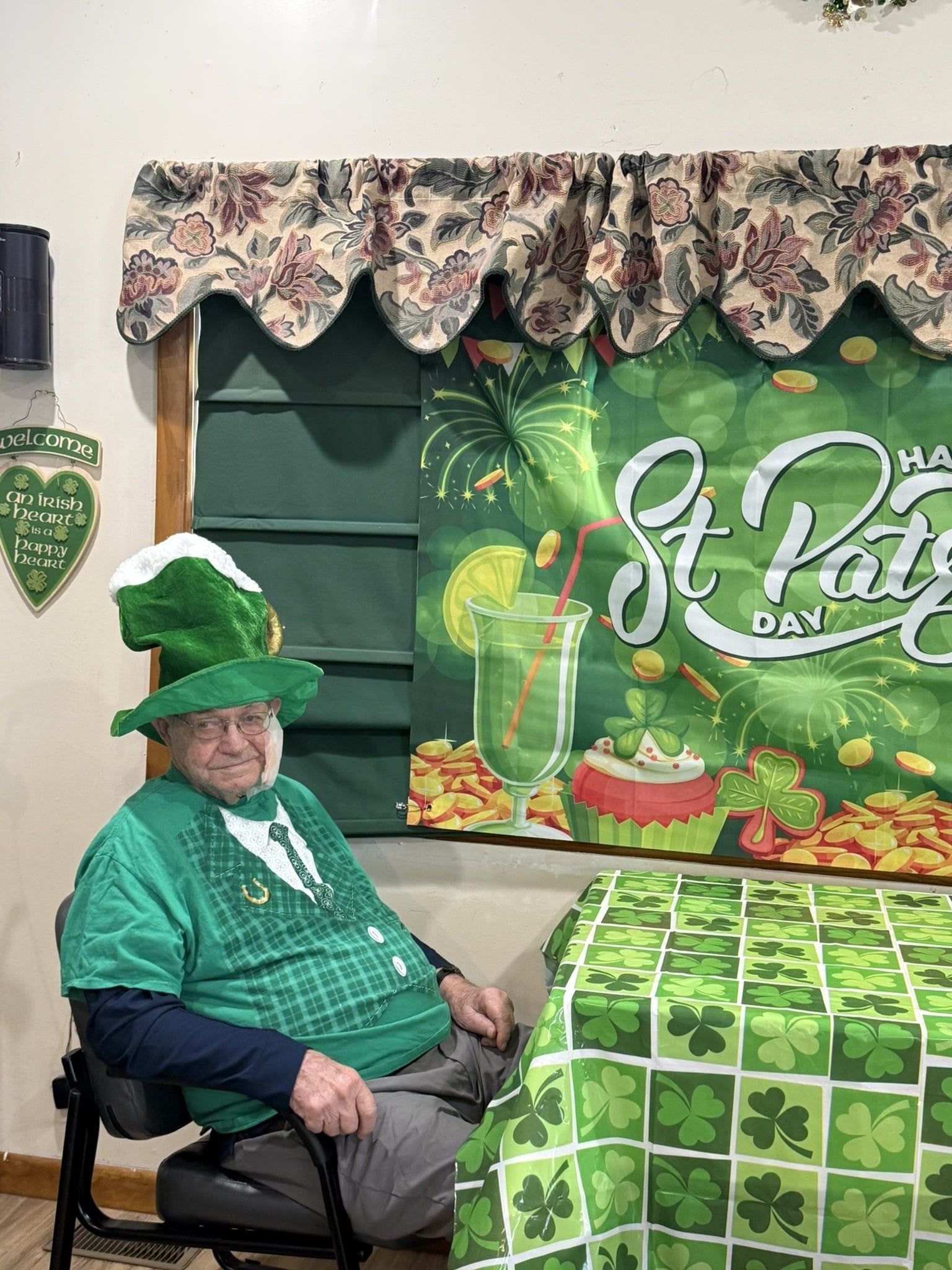 A person wearing a green leprechaun hat and shirt sits at a table with a shamrock-patterned cloth in front of festive decor.