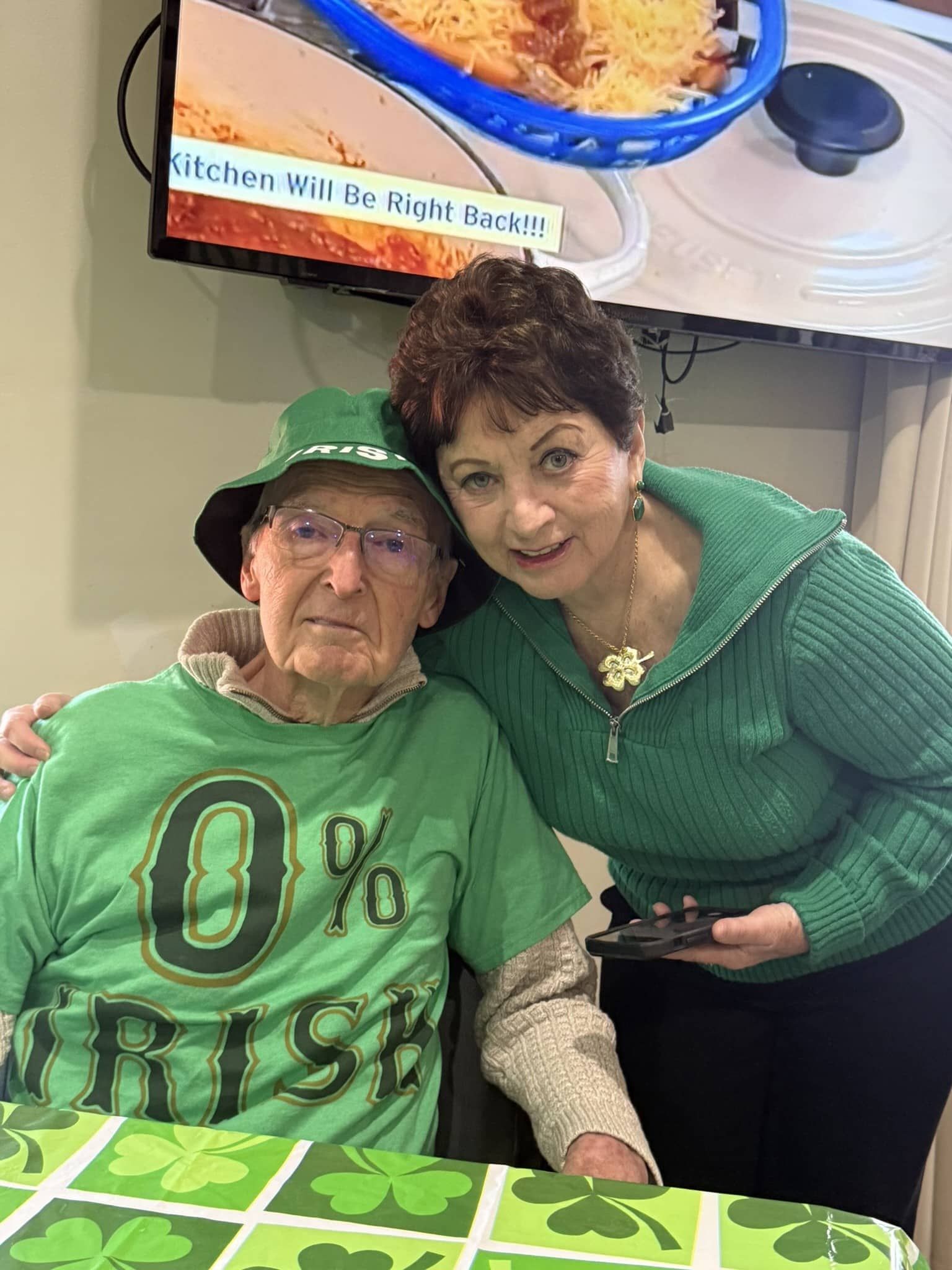 Two people wearing green clothing and accessories pose together indoors at a table with a shamrock-patterned tablecloth.