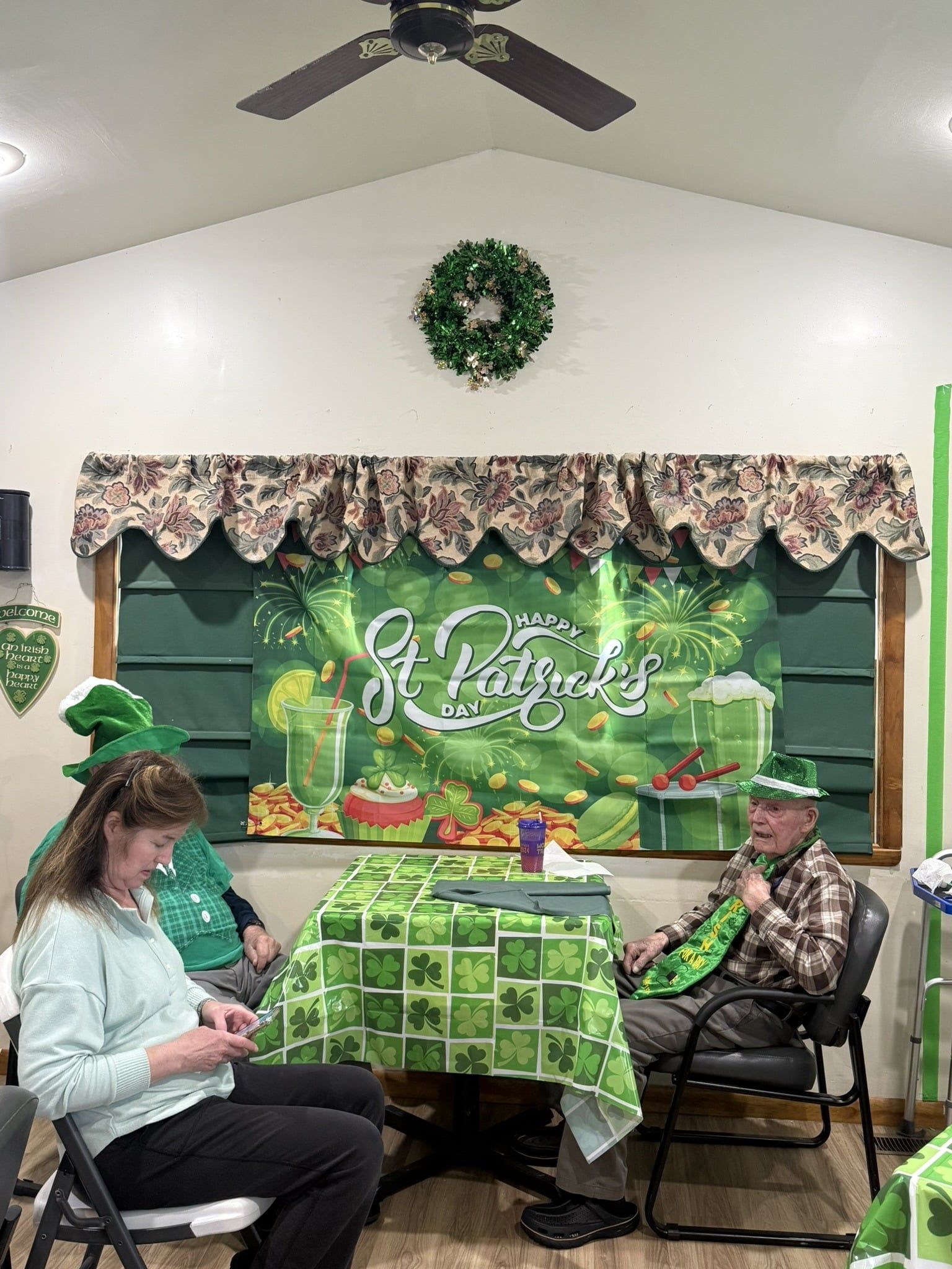 Two people in festive green St. Patrick's Day attire sit at a table decorated with a shamrock print cloth in a home setting.