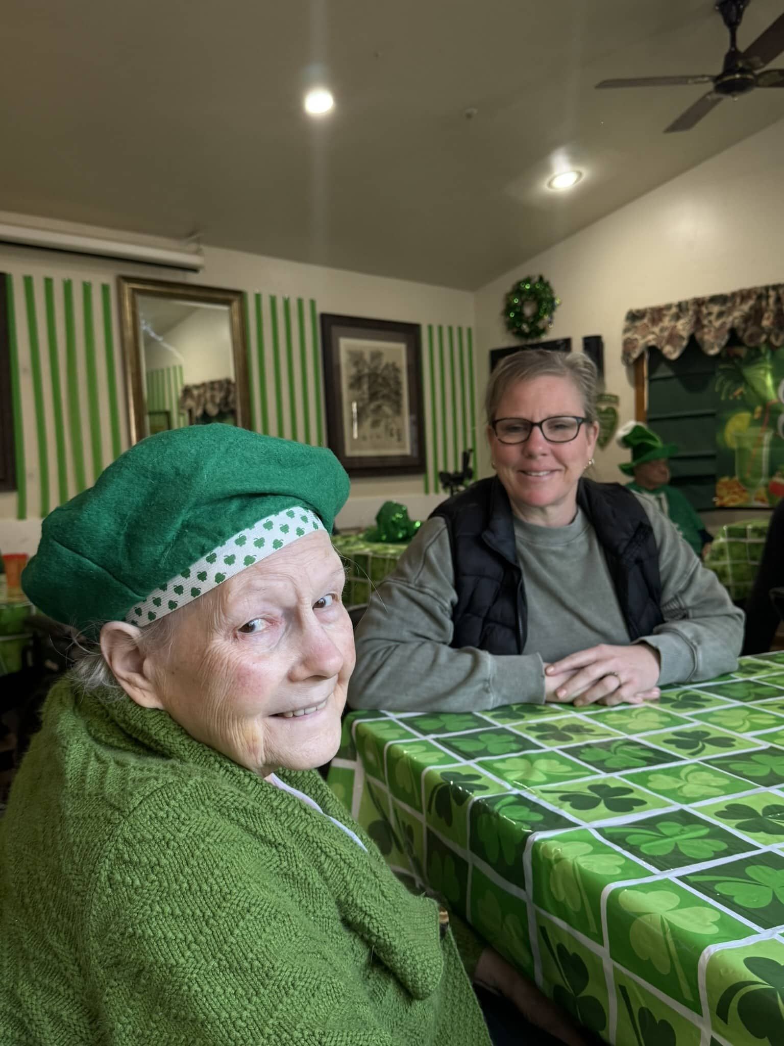 Two people in green outfits sit at a table with a shamrock-patterned tablecloth in a room decorated for St. Patrick's Day.