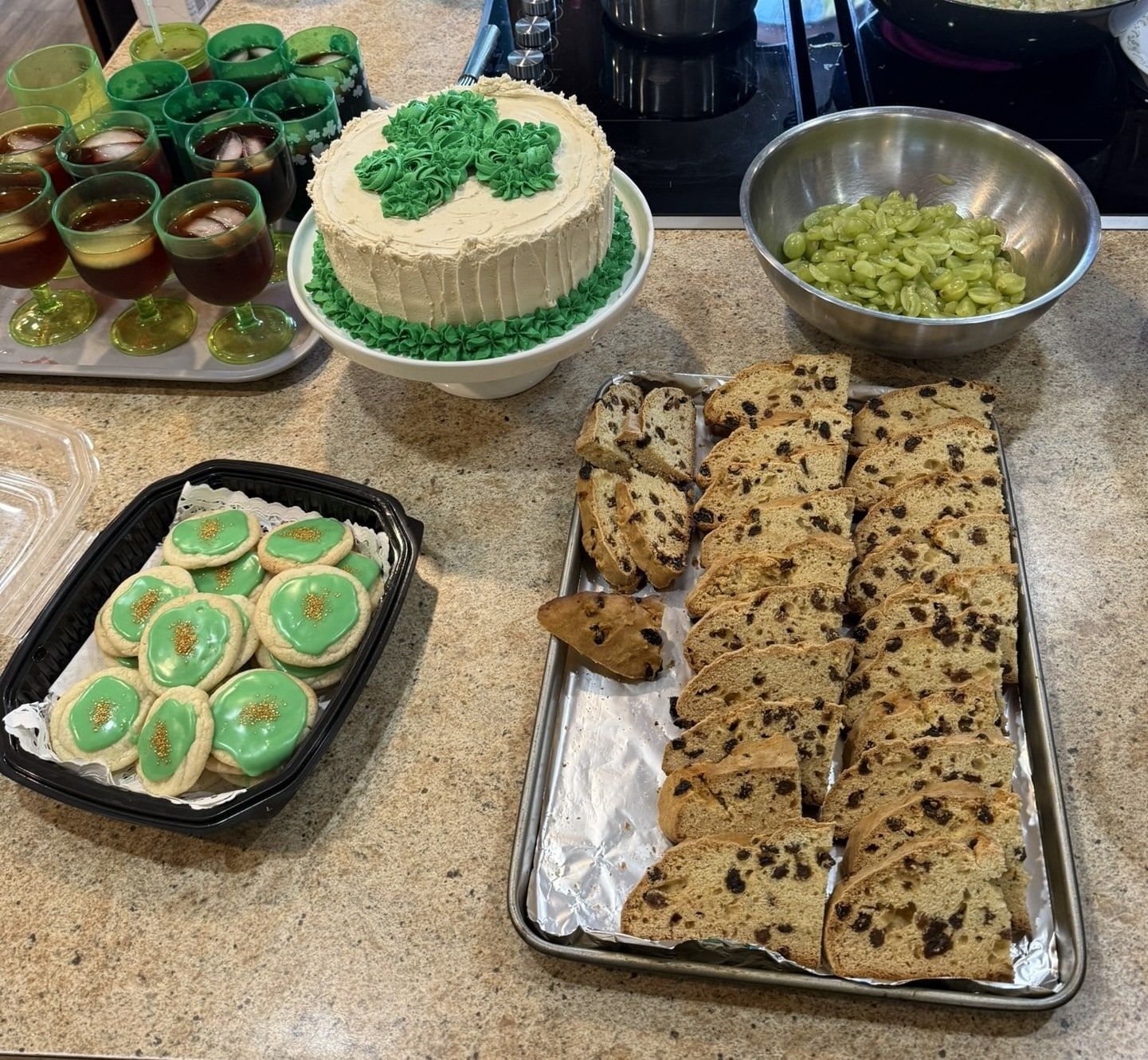 A St. Patrick’s Day spread featuring a shamrock cake, frosted cookies, slices of soda bread, grapes, and green drinks.