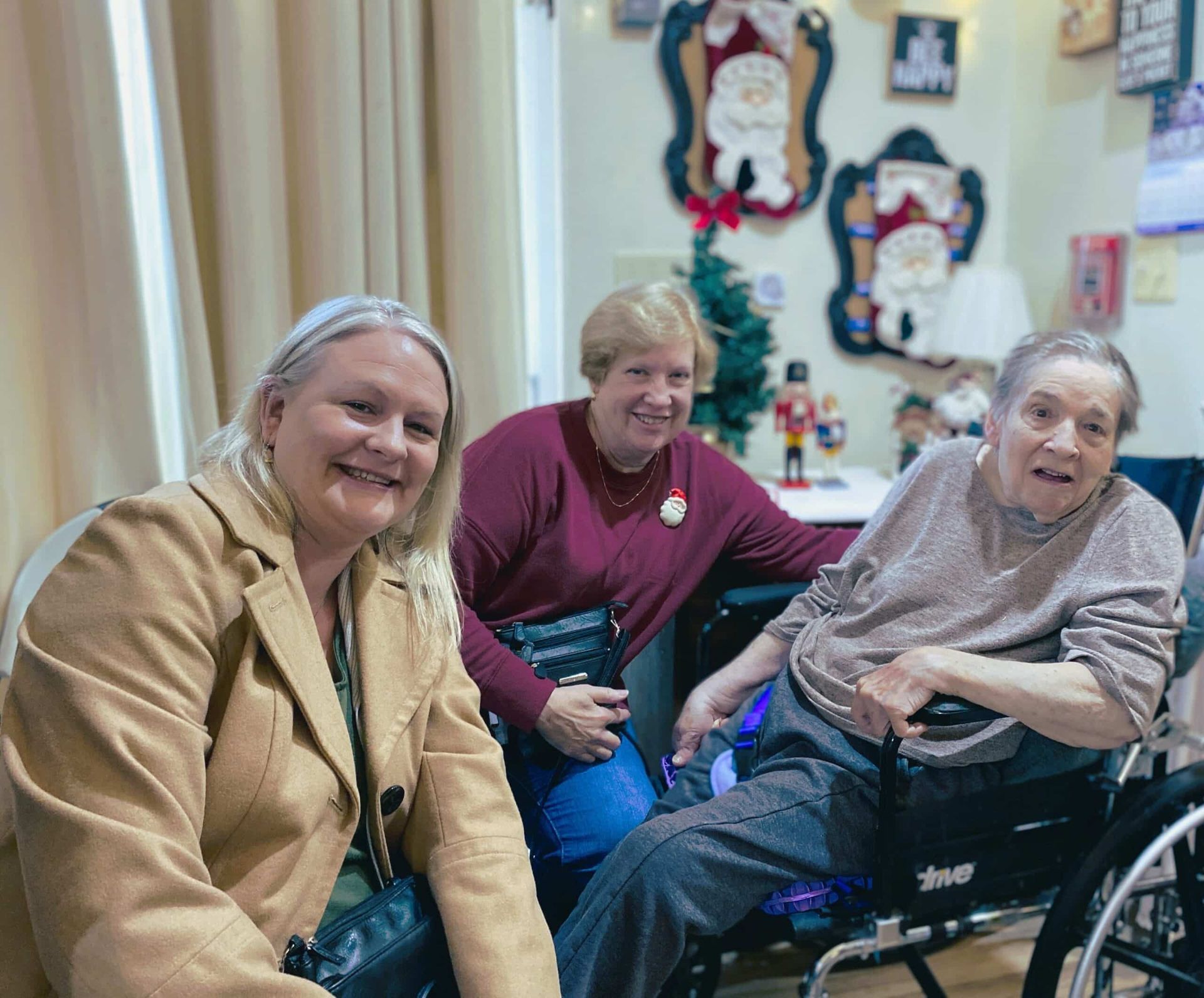 Three smiling people pose together indoors, with one person seated in a wheelchair in a room decorated for the holidays.
