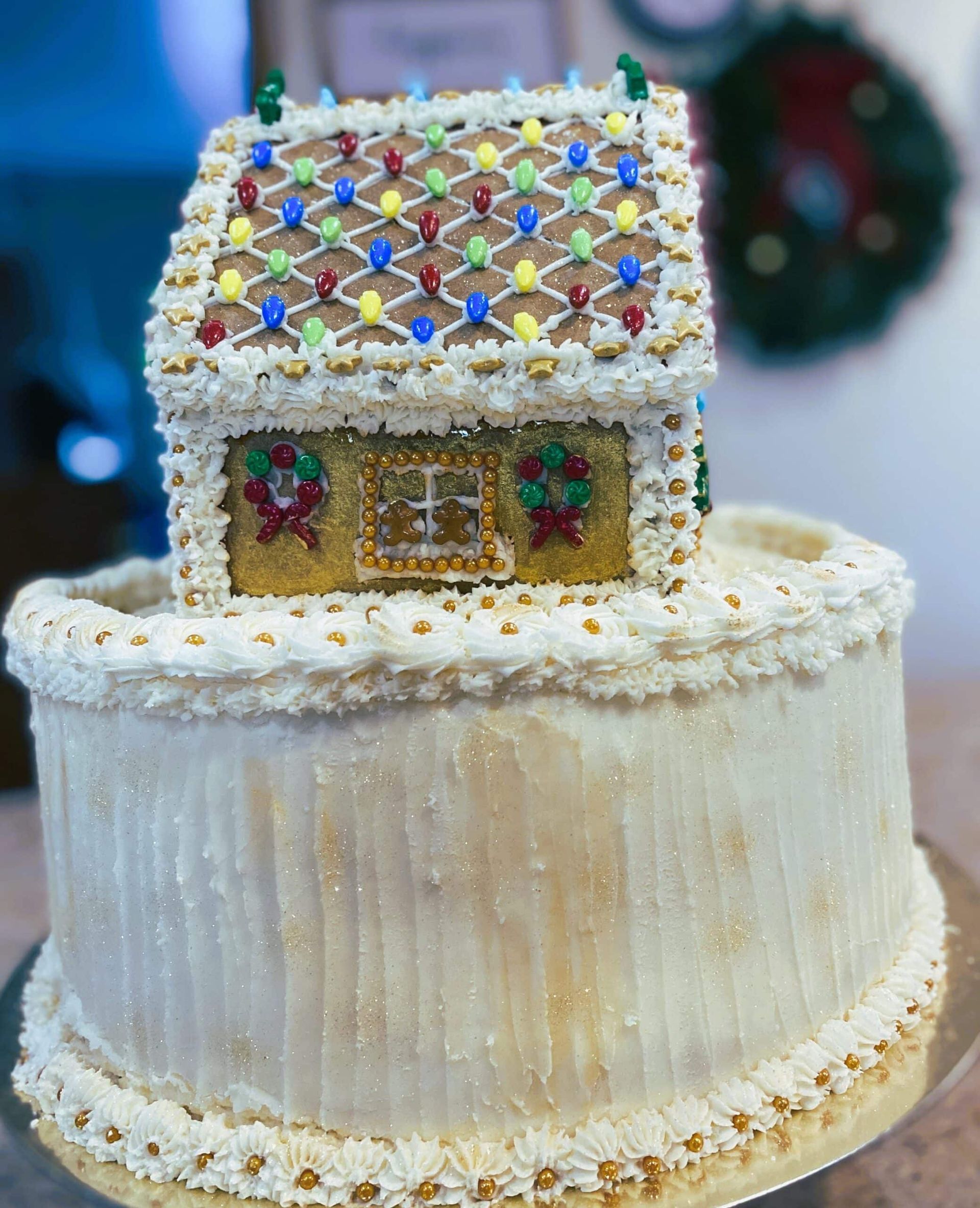A gingerbread house with a light-dotted roof sits atop a tall, white frosted cake decorated with golden accents.