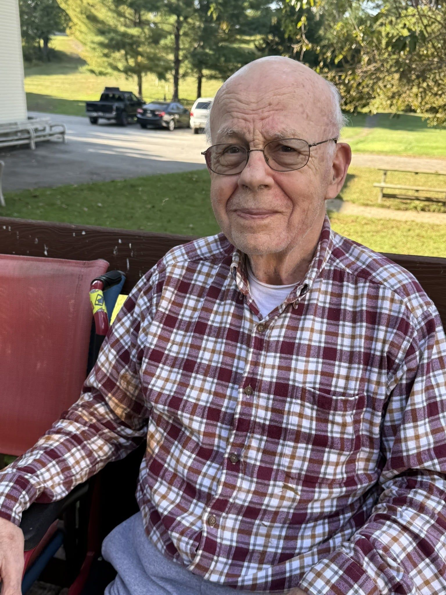 A person wearing a plaid shirt and glasses sits on a porch, smiling faintly with a grassy area and vehicles in the back.