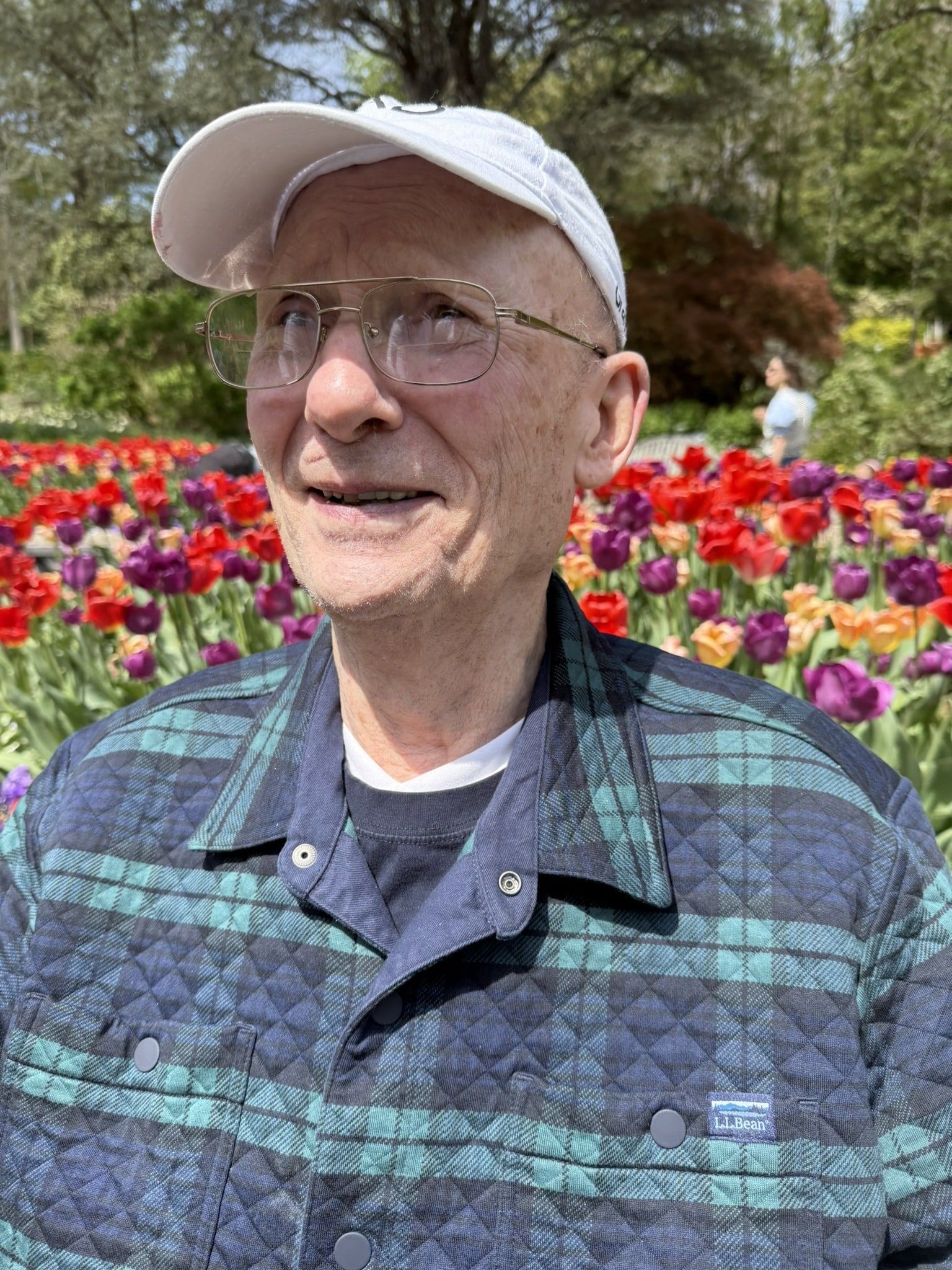 A person wearing a white baseball cap, glasses, and a quilted plaid jacket stands in front of a field of colorful tulips.