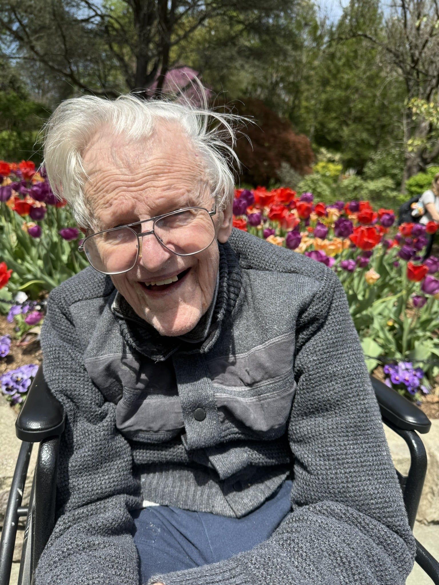 A smiling person in a gray sweater and wheelchair sits outside in front of a colorful garden of blooming tulips.