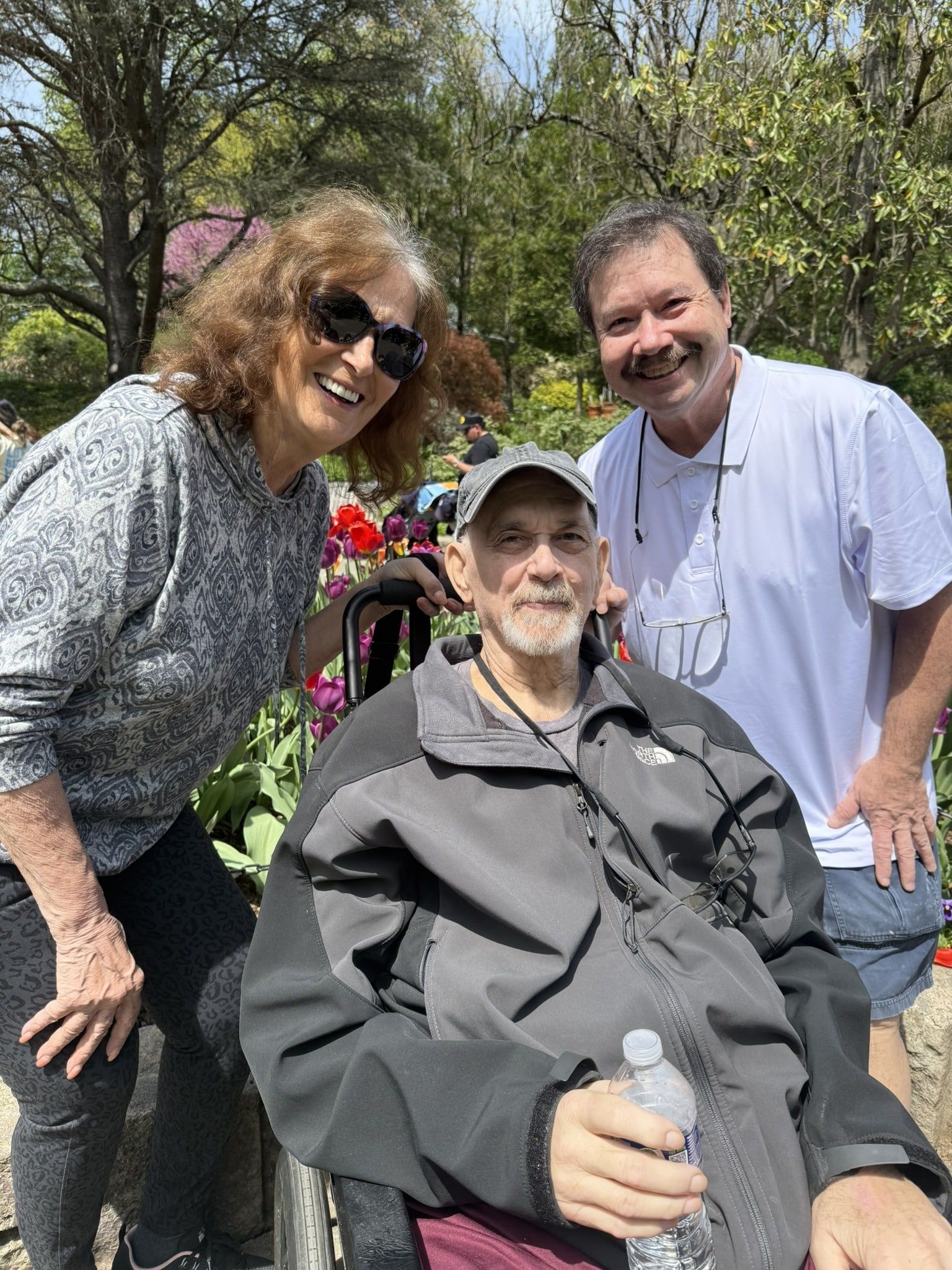 Three people smile together in an outdoor, garden-like setting. One person sits in a wheelchair in the foreground.