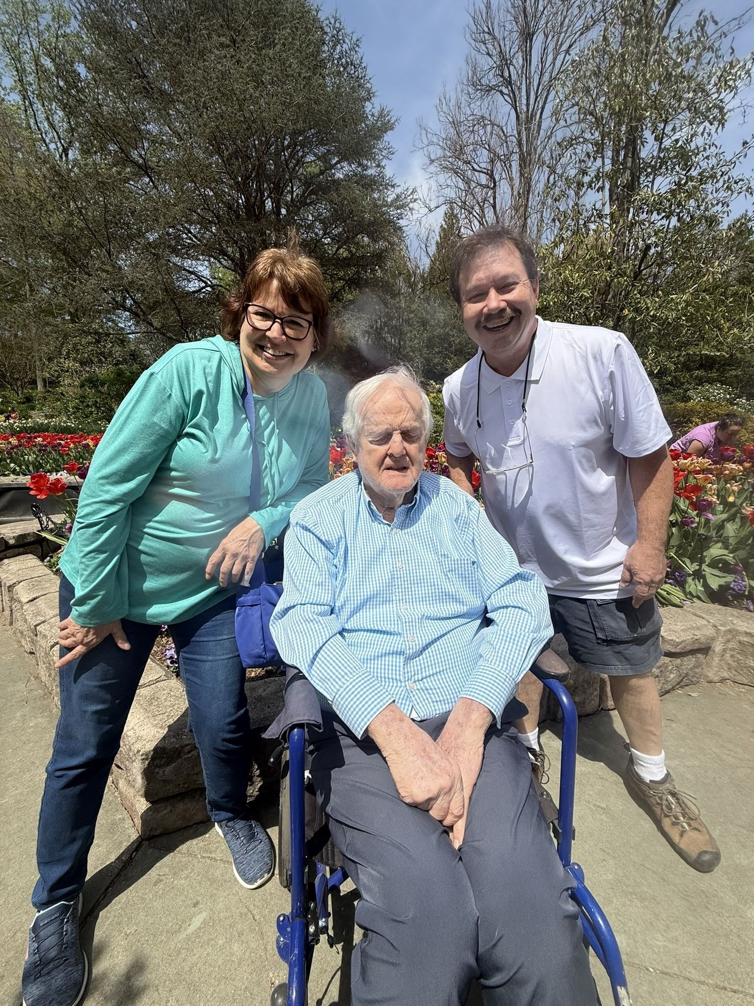 Three smiling people pose outdoors in a sunny garden with red flowers; one person sits in a blue wheelchair.